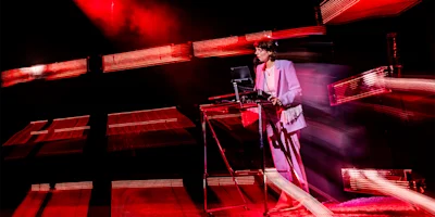 A person in a light-colored suit performs music on stage using electronic equipment, surrounded by dynamic red lighting effects.