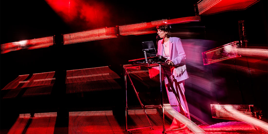 A person in a light-colored suit performs music on stage using electronic equipment, surrounded by dynamic red lighting effects.