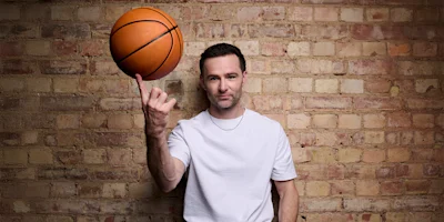A man in a white t-shirt spins a basketball on his index finger while standing in front of a brick wall.