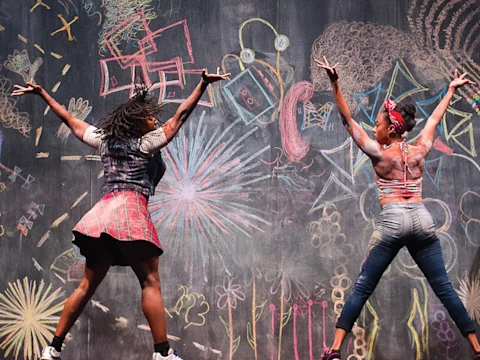 Two people with arms raised stand in front of a large chalkboard filled with colorful, abstract chalk drawings and patterns.