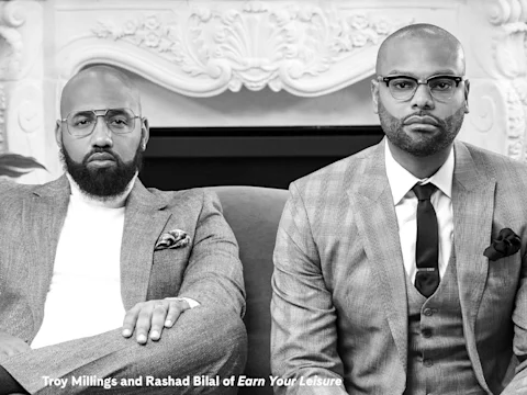Two men in suits sit on a couch in front of an ornate fireplace, looking directly at the camera. Text reads: "Troy Millings and Rashad Bilal of Earn Your Leisure.