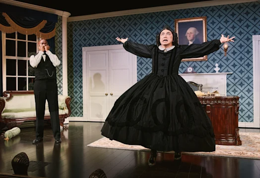 A performer in a black mourning dress flings both arms wide mid-outburst while another watches from a window.