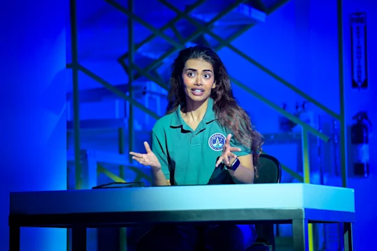 A woman in a green shirt sits at a table, gesturing with her hands, under blue lighting with industrial shelves and fire extinguishers in the background.