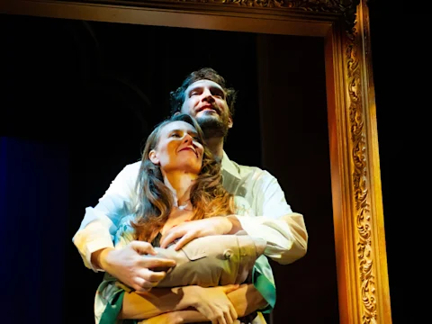 A man stands behind a woman with his arms around her waist, both looking upward, framed by an ornate gold frame on stage.