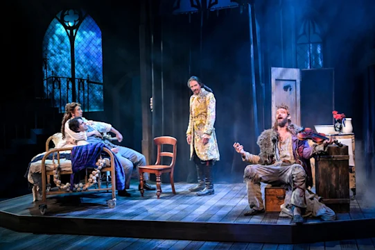 Four actors perform in a stage production of A Christmas Carol, featuring a man playing the violin while seated on a trunk.