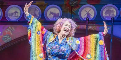 A woman with curly hair smiles and raises her arms on stage, wearing a colorful, rainbow-patterned shawl and a floral dress, with circular lights and images in the background.