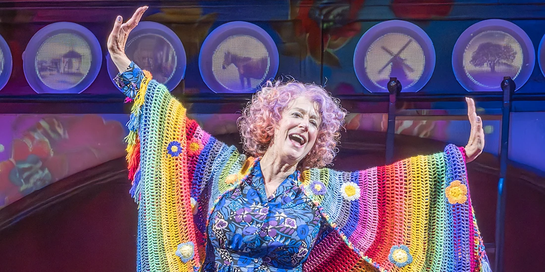 A woman with curly hair smiles and raises her arms on stage, wearing a colorful, rainbow-patterned shawl and a floral dress, with circular lights and images in the background.