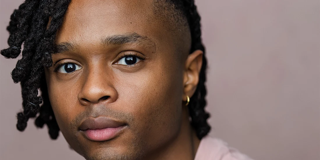 Close-up portrait of a person with medium brown skin, short twisted hair on one side, wearing a small gold hoop earring, and looking at the camera against a neutral background.