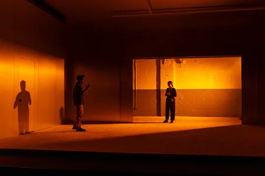 Two people stand apart in a dimly lit, orange-hued room; one is near the back wall, the other closer to the foreground, with their shadow cast on the wall.