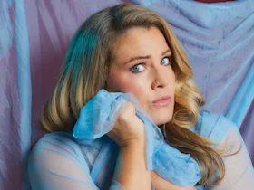 A woman with long blonde hair holds a piece of sheer blue fabric to her cheek, looking slightly upward with a neutral expression. Blue drapery is in the background.