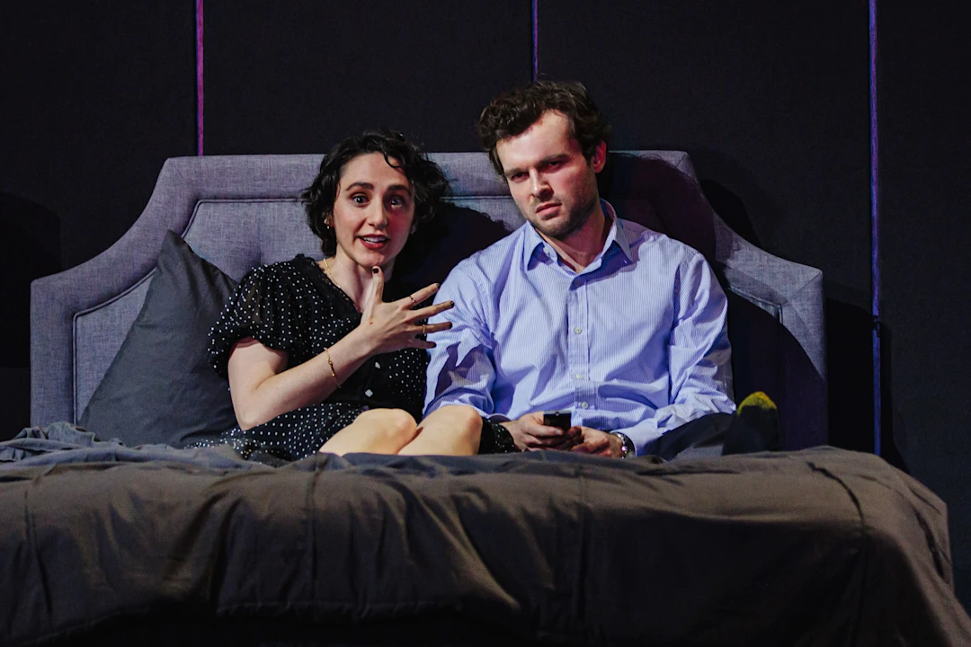 Two people sit on a bed; the woman gestures while speaking, and the man, holding a remote control, looks forward with a serious expression.