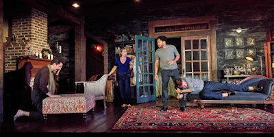Four actors perform an intense scene on a set resembling a cozy, rustic living room with vintage furniture and a patterned rug.