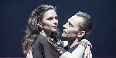 Two actors on stage, both wearing microphones, embrace closely while looking at each other under dramatic lighting.