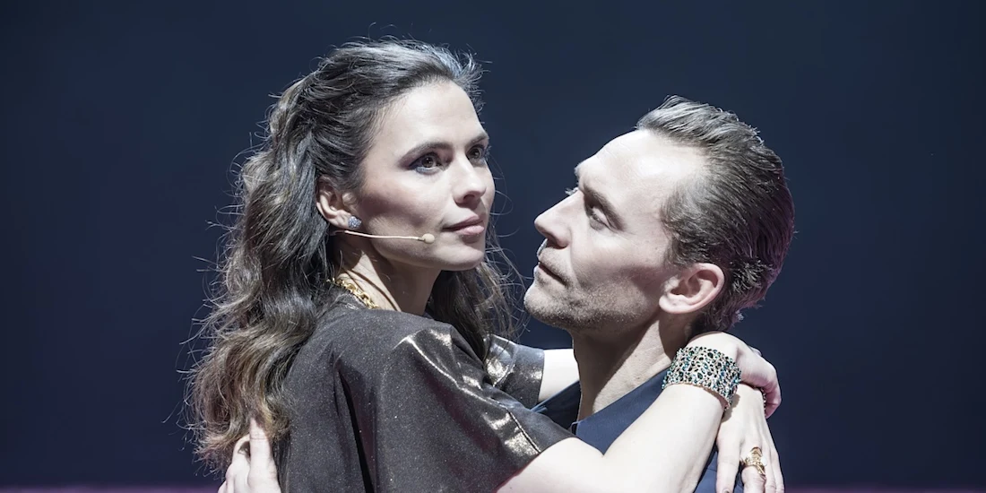 Two actors on stage, both wearing microphones, embrace closely while looking at each other under dramatic lighting.