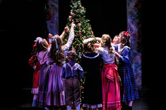 Children in festive attire gather around a decorated Christmas tree, reaching to hang ornaments on its branches.