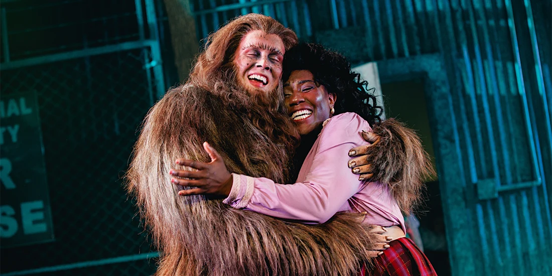 A person in a furry costume hugs another person wearing a pink shirt and red skirt; both are smiling and standing in front of a metal fence.