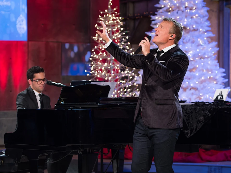 A man sings into a microphone while another man plays a grand piano. Decorated Christmas trees and festive lighting are visible in the background.