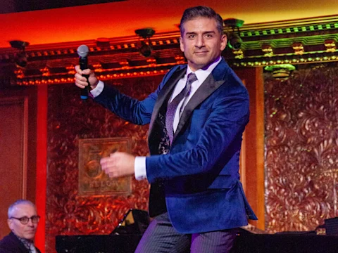 A man in a blue velvet suit jacket holds a microphone and performs on stage; another man is seated at a piano in the background.