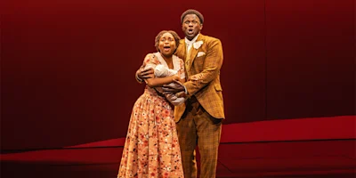 Two actors on stage sing together; the woman wears a floral dress and holds a bundled baby, while the man wears a brown plaid suit, both standing against a red background.