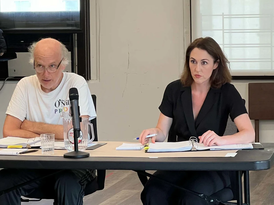 Two people are seated at a table with documents and a microphone in front of them, engaging in a serious discussion.