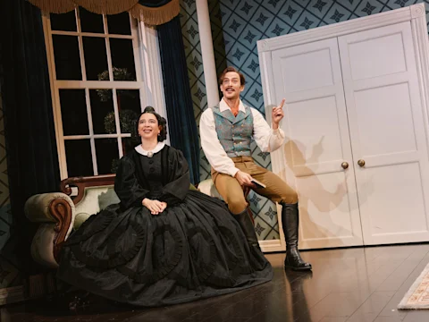 Two actors in period costumes perform on stage; the woman sits on a green sofa in a black dress, while the man gestures with one finger raised, sitting near a white wardrobe.