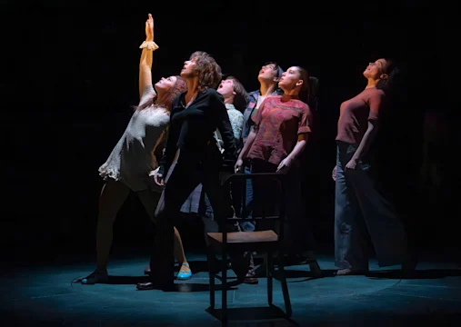Six people stand closely together on a dimly lit stage, with one person reaching upward. A chair is placed in front of them.