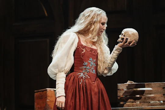 A woman in a red Renaissance-style dress holds a skull in one hand, standing on stage in front of a dark wooden background.