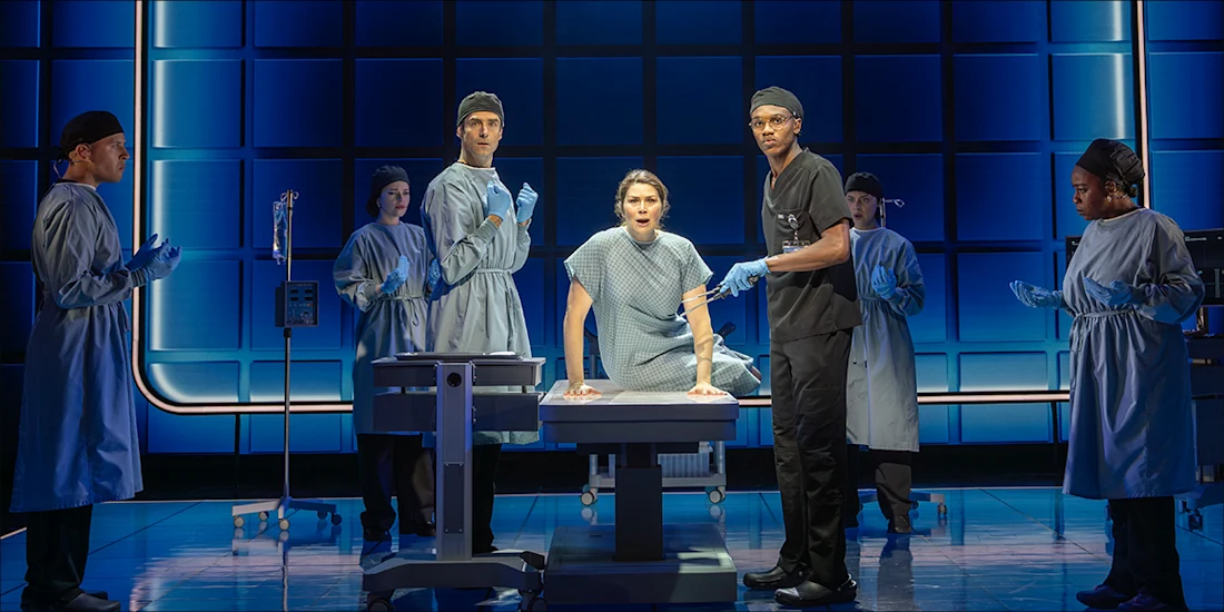 A patient in a hospital gown sits on an exam table surrounded by six medical professionals in scrubs and blue gloves on a stage with a blue grid background.