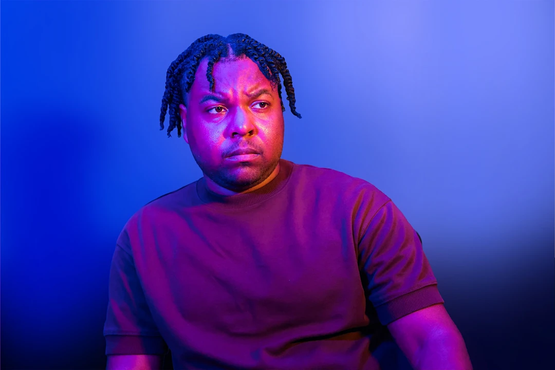 A man with braided hair and a serious expression sits against a blue and purple gradient background.