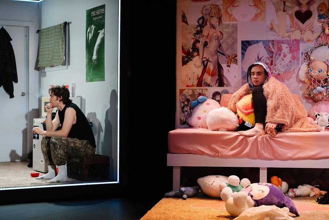 Two people in a bedroom: one sits on a bed surrounded by stuffed toys and anime posters, wearing a pink robe; the other sits in a separate area, looking thoughtful.