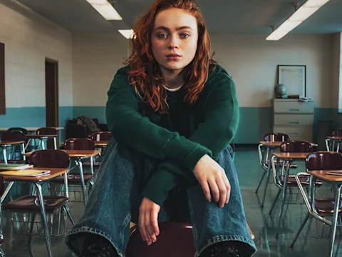 Person with red hair sits alone in an empty classroom, wearing a green long-sleeve shirt and blue jeans, looking directly at the camera.