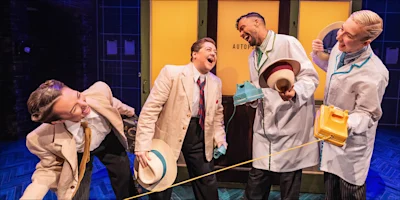 Four people in suits and lab coats laugh and interact on stage, holding hats and yellow and blue phones, with a brightly lit backdrop behind them.