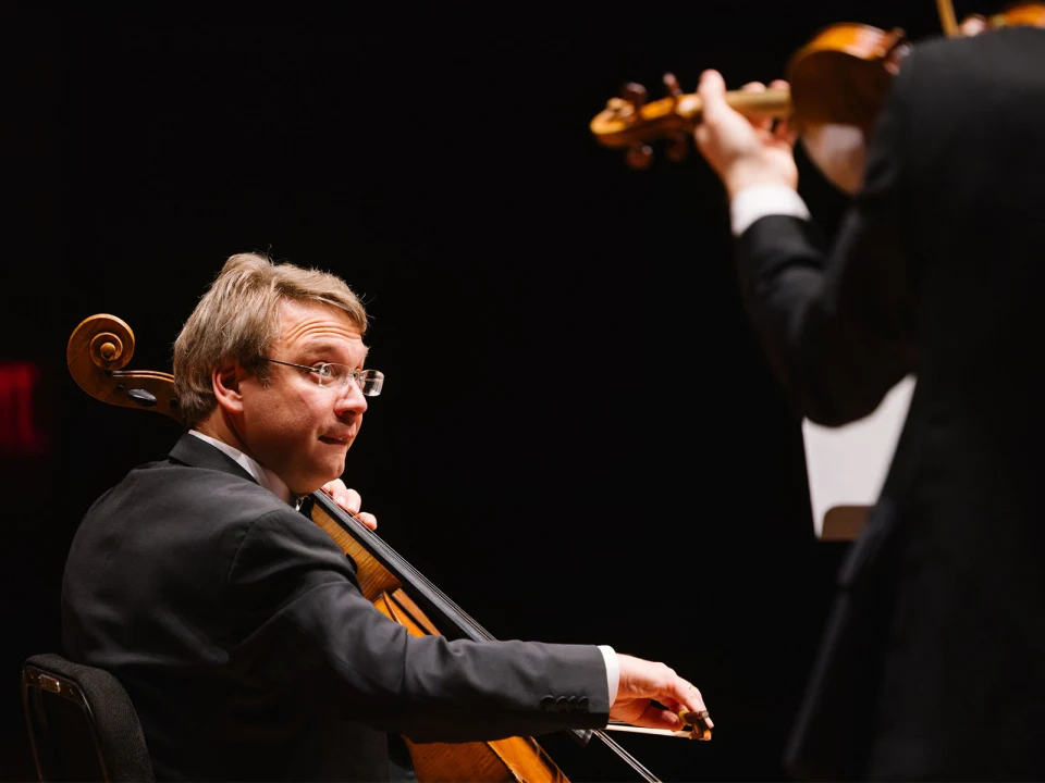 A cellist in a formal suit performs on stage while another musician plays the violin in the foreground, partially out of focus.