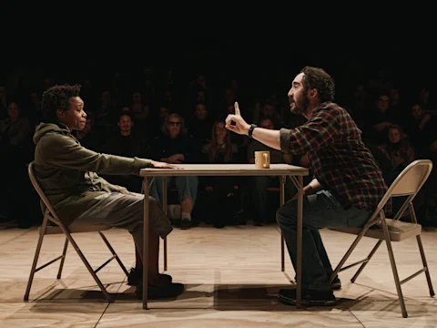 Two people sit across from each other at a table, engaged in an intense conversation, with an audience watching in the background.