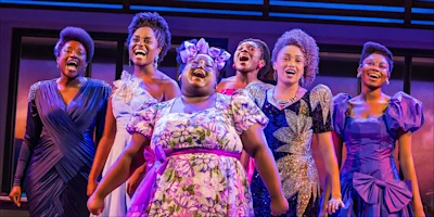 Six women in colorful, formal dresses sing together on stage under bright lighting.