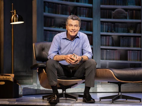 A man in a blue shirt sits on a chair in front of a bookshelf, smiling. A floor lamp is beside him and a box labeled "Psychology" is partially visible.