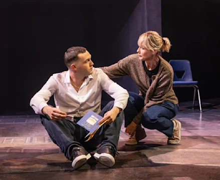 A man in a white shirt sits on the floor holding papers, while a woman with a ponytail kneels beside him, resting her hand on his shoulder. An empty chair is in the background.
