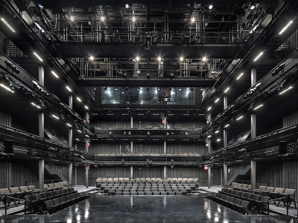 Interior view of a modern theater with tiered seating, black walls, and exposed lighting rigs overhead, photographed from the stage facing the audience.