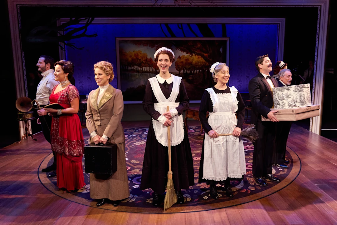 Six actors in early 20th-century costumes stand in a line on stage, with household props, against a backdrop of a framed painting and wallpaper.