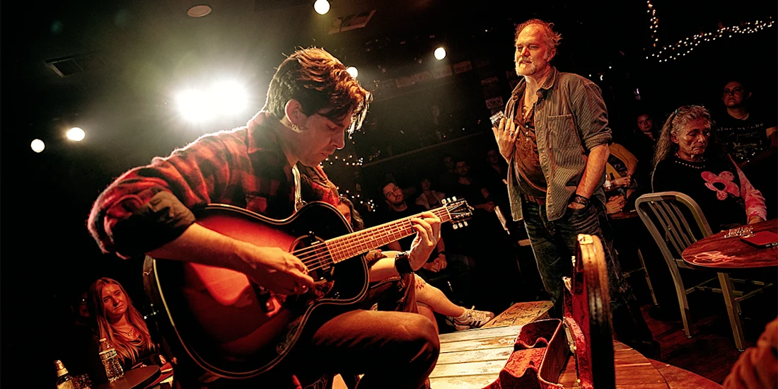 A musician plays an acoustic guitar onstage in a dimly lit venue as an audience watches. Some people sit at tables, while one person stands nearby.