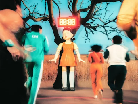 People in colorful outfits run toward a large doll with a timer above it, set in an outdoor area with a tree in the background.