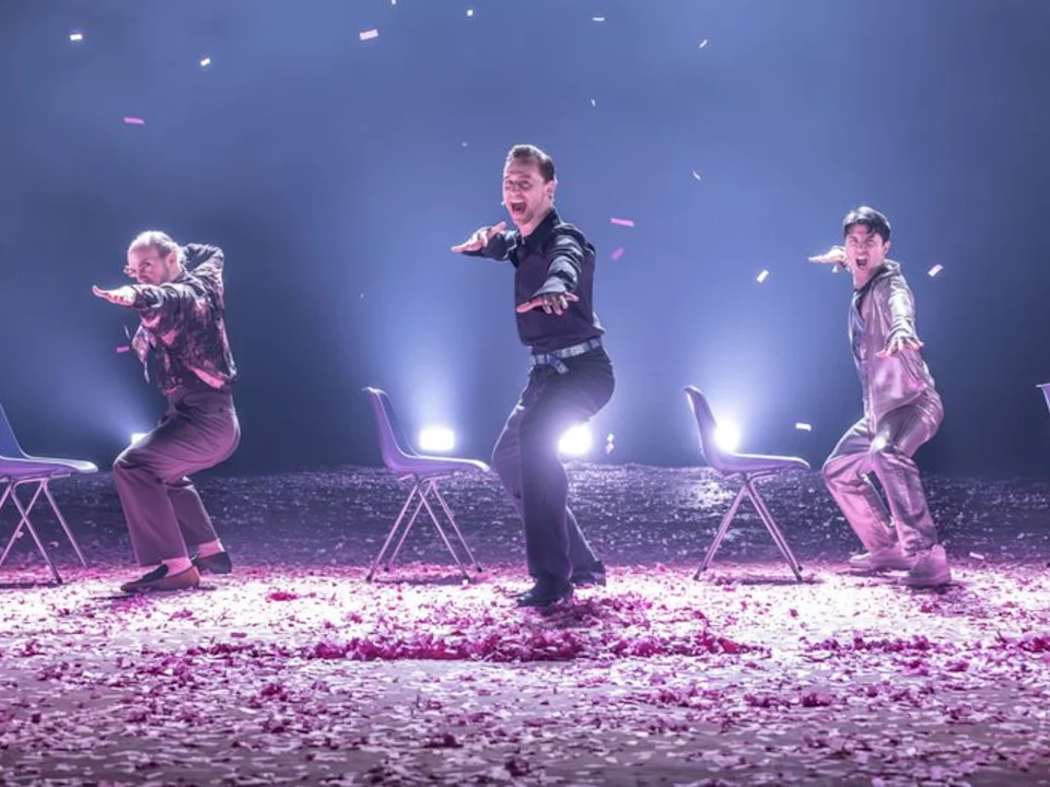 Three people pose energetically in front of chairs on a stage covered with flower petals, lit by bright spotlights and surrounded by falling confetti.