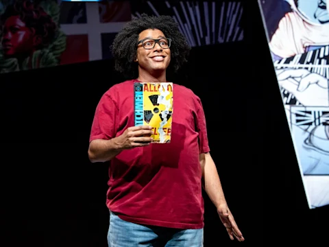 A person with curly hair and glasses, wearing a red shirt, stands on stage holding a comic book. Comic-themed artwork is visible in the background.