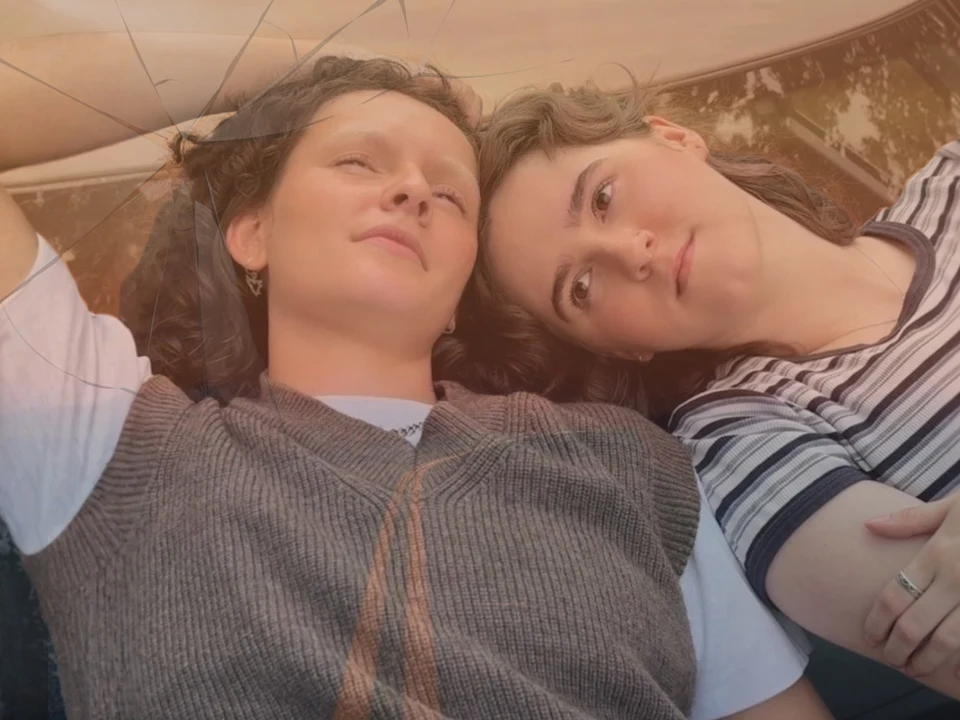 Two people lie side by side, one looking upward and the other gazing at them, with relaxed expressions and a cracked glass surface in the foreground.