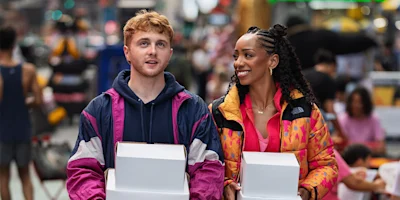 Two people walk outdoors holding white boxes, dressed in colorful jackets. A busy, blurred crowd and city setting are visible in the background.