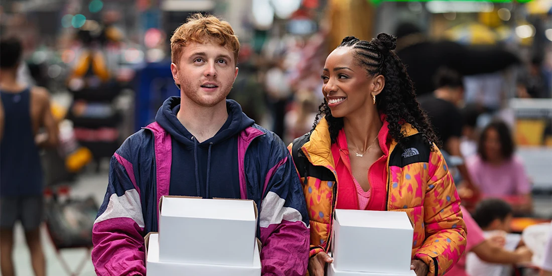 Two people walk outdoors holding white boxes, dressed in colorful jackets. A busy, blurred crowd and city setting are visible in the background.