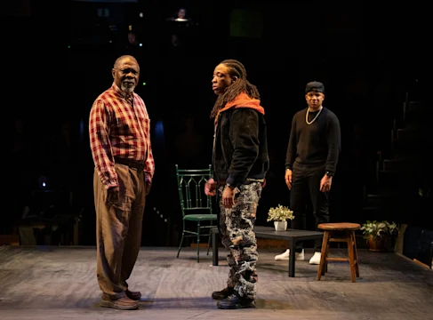 Three people stand on a stage with minimal props, including chairs, a stool, and potted plants, under stage lighting in a dark theater setting.