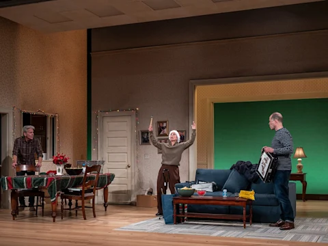 Three adults are in a living room; one gestures excitedly with arms raised while the others watch. The setting includes a sofa, a dining table, and festive lights.