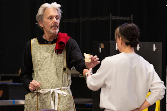 An older man in a costume apron hands an object to a woman in casual clothing. They appear to be interacting indoors, possibly during a rehearsal or demonstration.