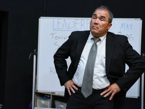 A man in a suit and tie stands with hands on hips in front of a whiteboard with text about leadership.